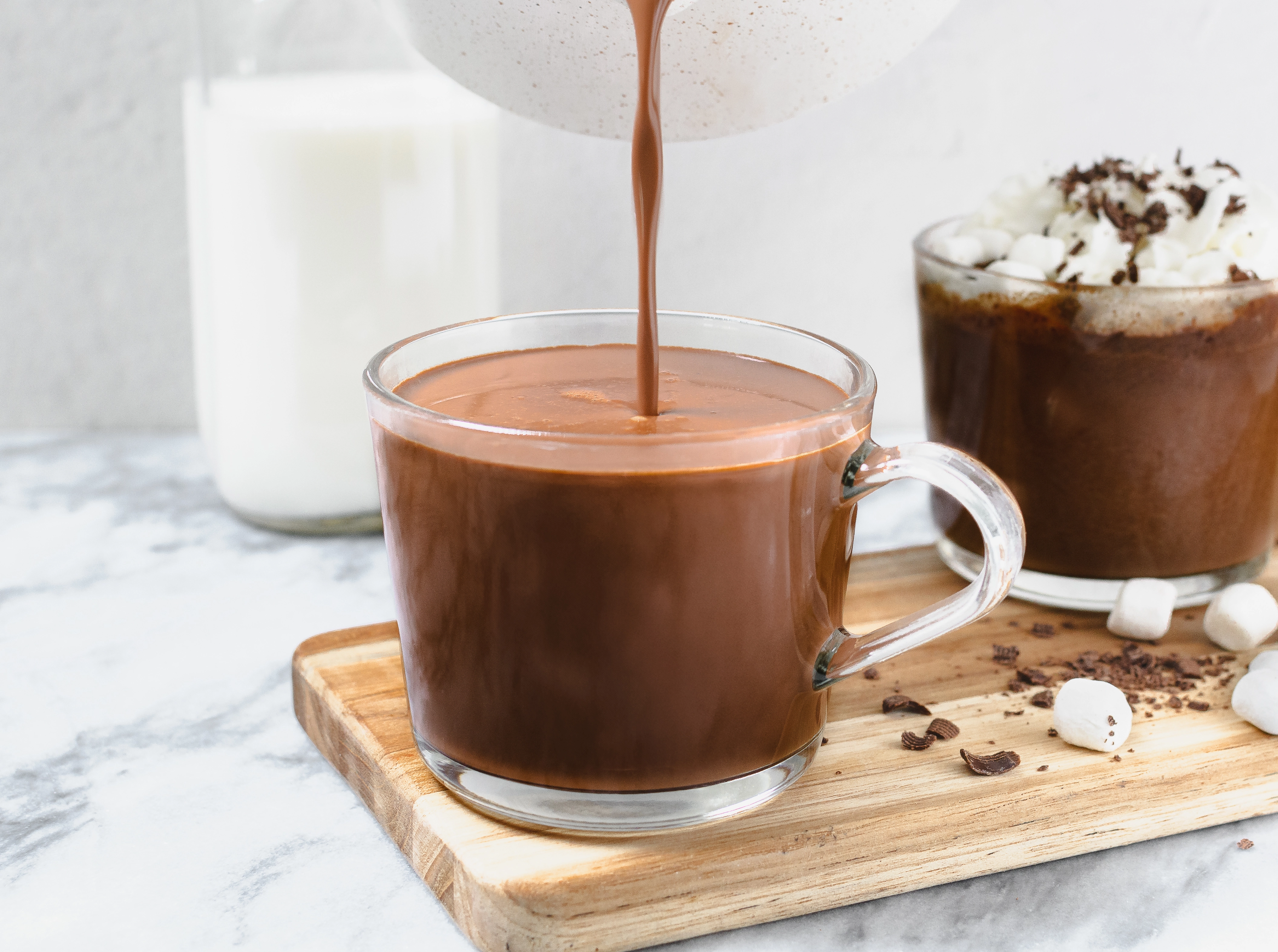 A glass mug of homemade cocoa with milk, alongside another mug topped with marshmallows and chocolate chips.