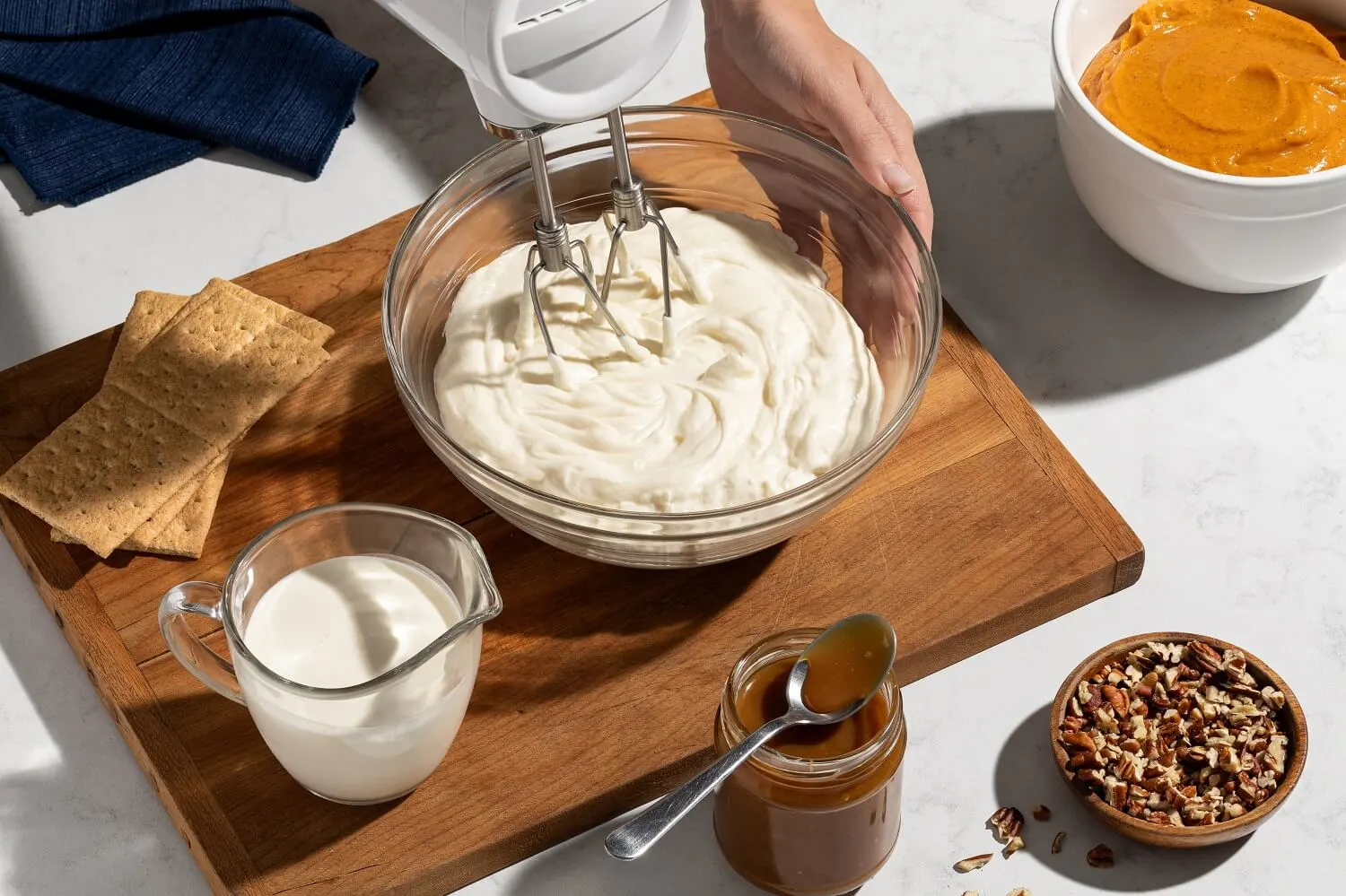 Hand-mixing cream cheese on a wooden table with graham crackers, a cup of milk, and a caramel jar in the background.