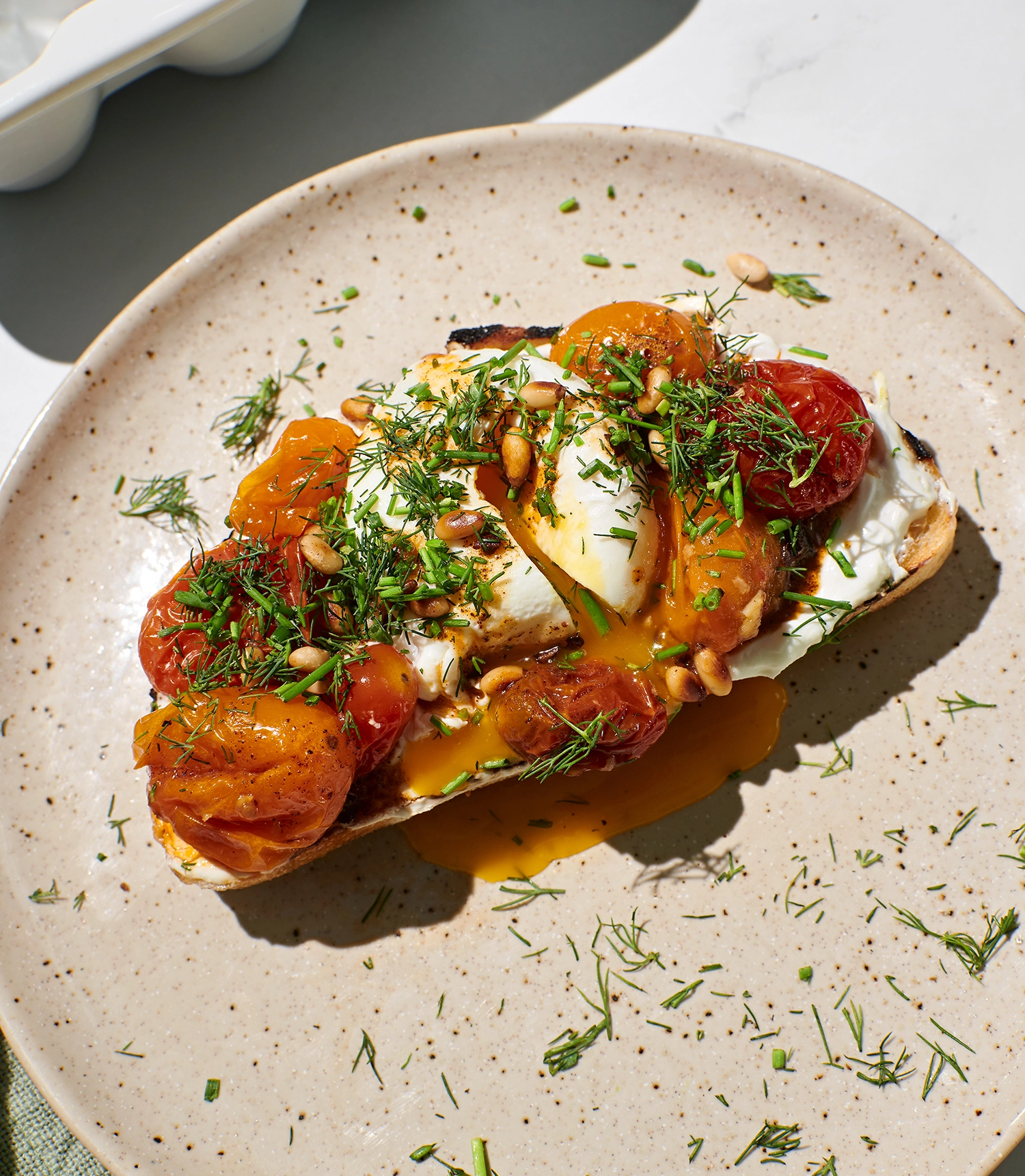 Garlicky Greek yogurt spread onto toast topped with roasted tomatoes, poached eggs, butter sauce, and garnished with toasted pine nuts, fresh chives, and dill served on a plate.