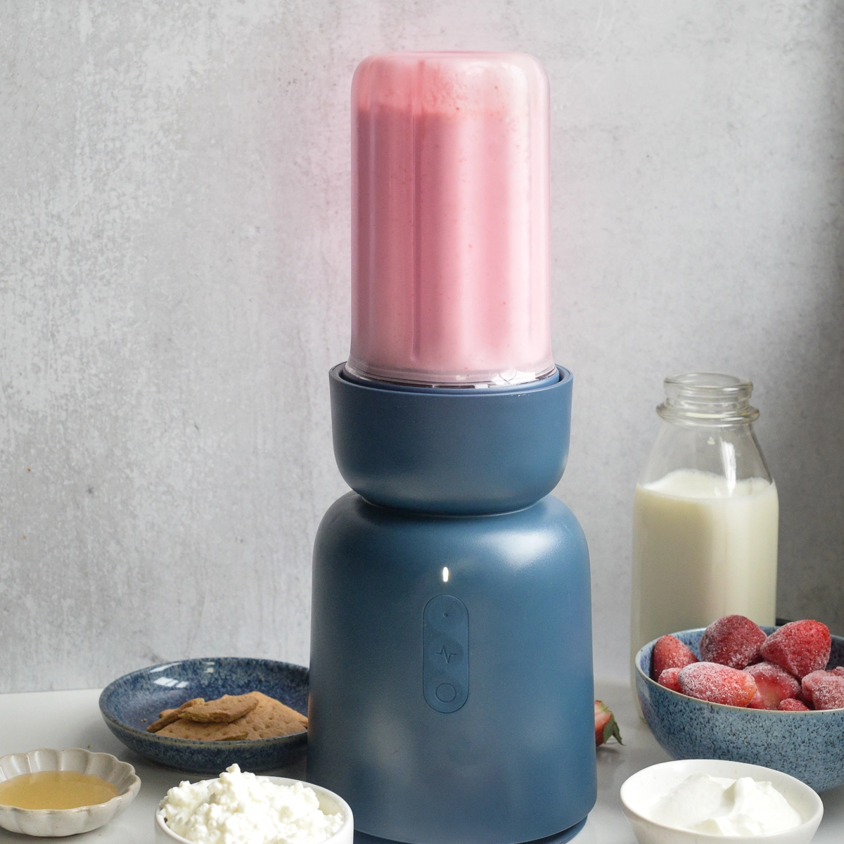 Strawberry cheesecake smoothie blended in blender, with ingredients in bowls beside blender