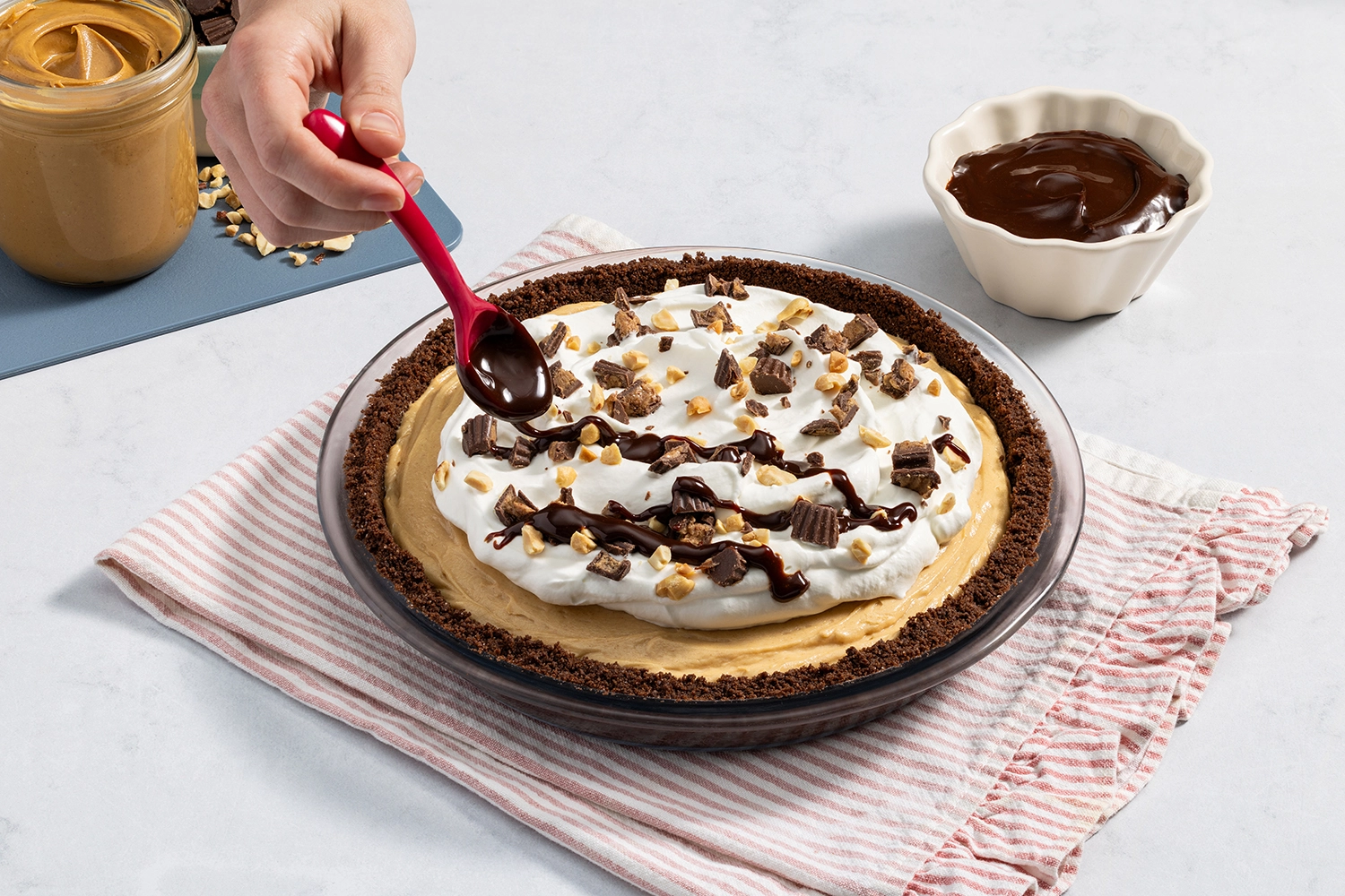 A hand using a spoon to drizzle chocolate sauce over a whipped-cream-topped peanut butter pie.