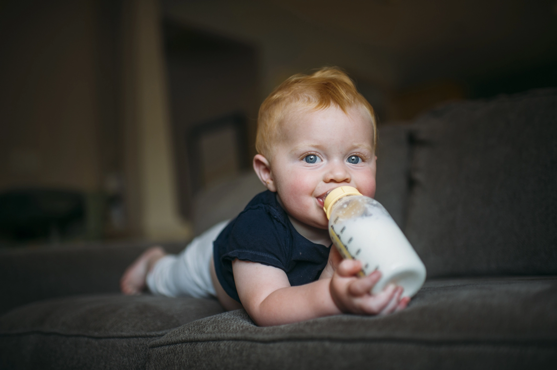 Happy baby laying on his stomach and drinking milk from a bottle on a couch.