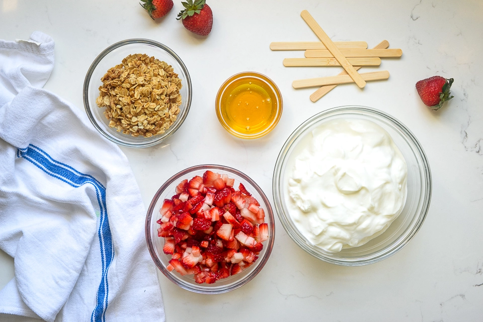 Ingredients for Greek yogurt popsicles: bowl of Greek yogurt, diced strawberries, granola, honey, and wooden sticks.