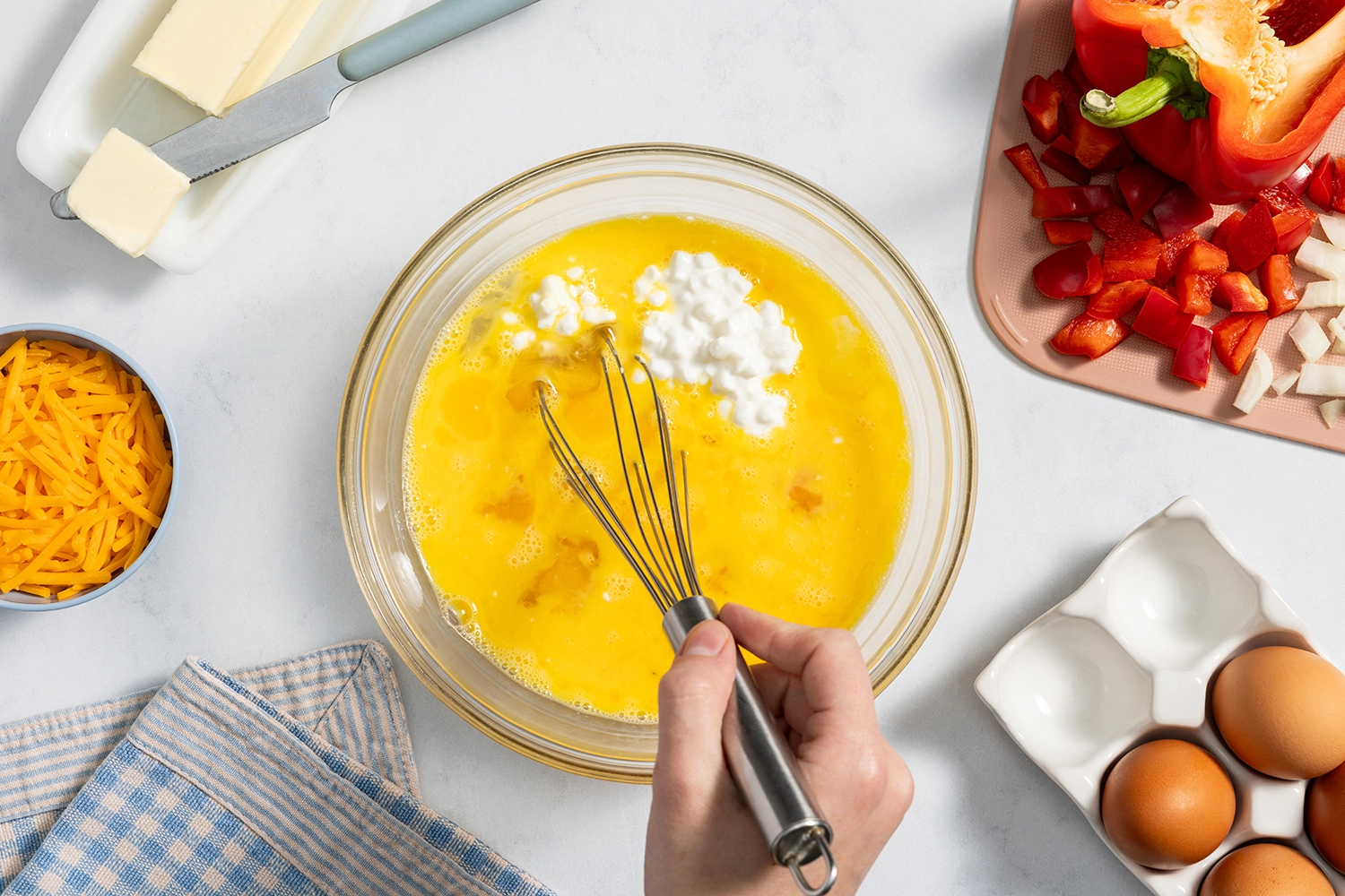 Overhead view of a hand whisking cottage cheese into raw eggs in a glass mixing bowl.