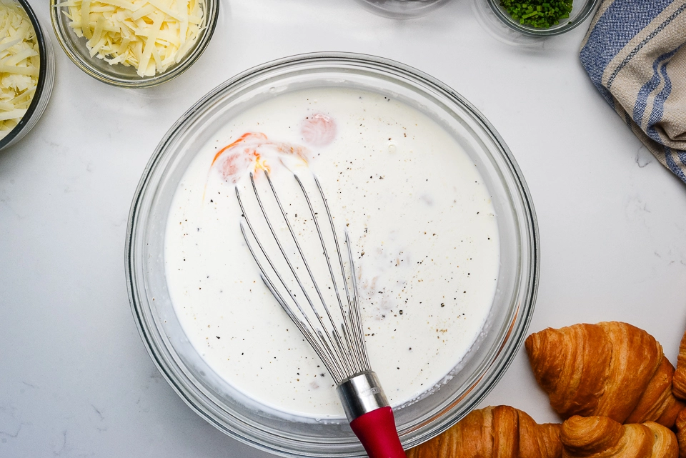Overhead view of a wire whisk mixing eggs and cream in a glass bowl.