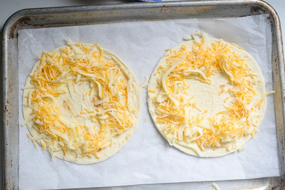 Two flour tortillas topped with a ring of shredded cheese on a parchment-lined baking sheet.