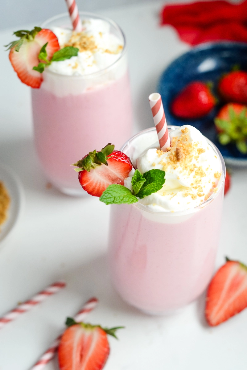 Strawberry cheesecake smoothie served in 2 glasses garnished with whipped cream, graham cracker crumbs and a fresh strawberry