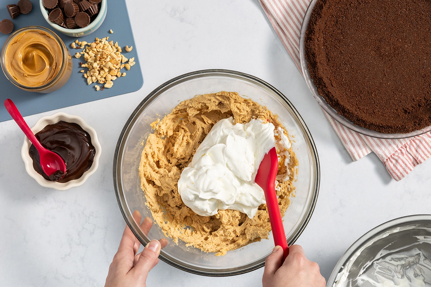Overhead view of hands using a spatula to fold fresh whipped cream into a peanut butter filling in a glass bowl.