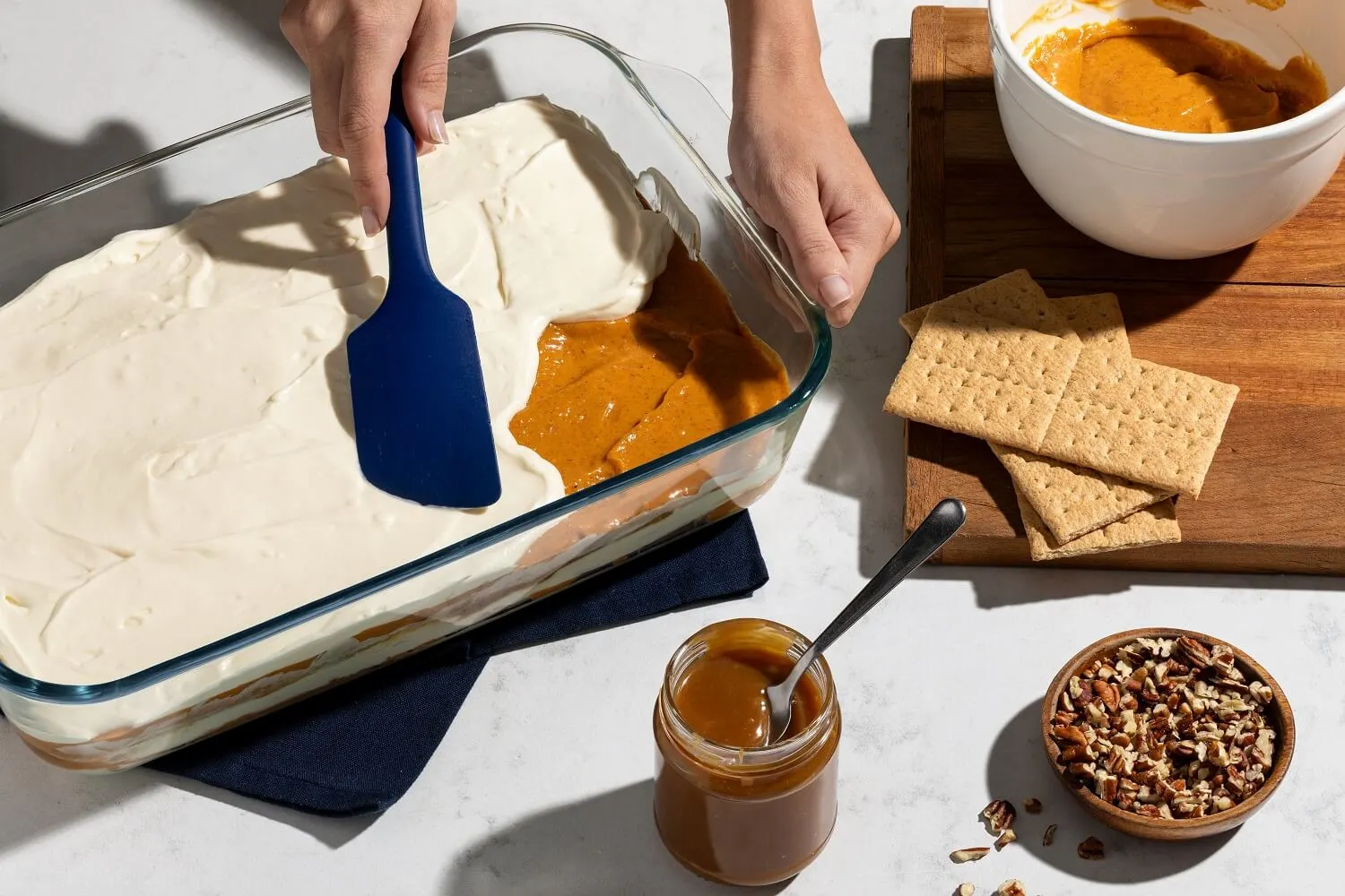 Two hands spreading cream cheese over caramel in a glass baking dish, with graham crackers, a jar of caramel, and a bowl of pecans.