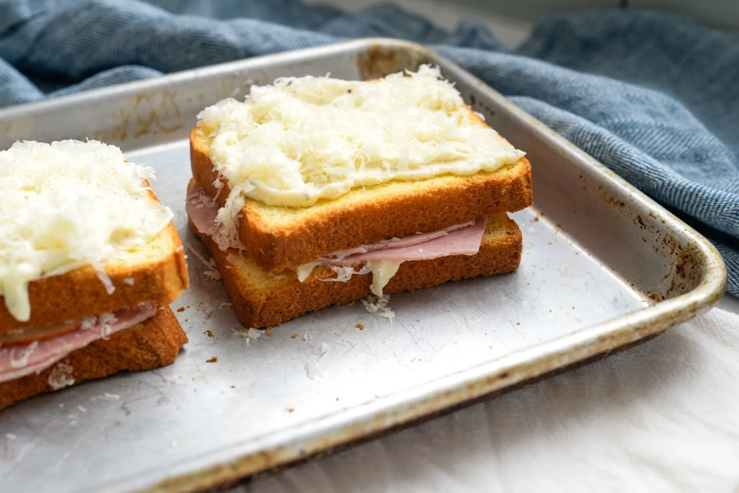 Two assembled Croque Monsieur sandwiches topped with béchamel in a baking pan.