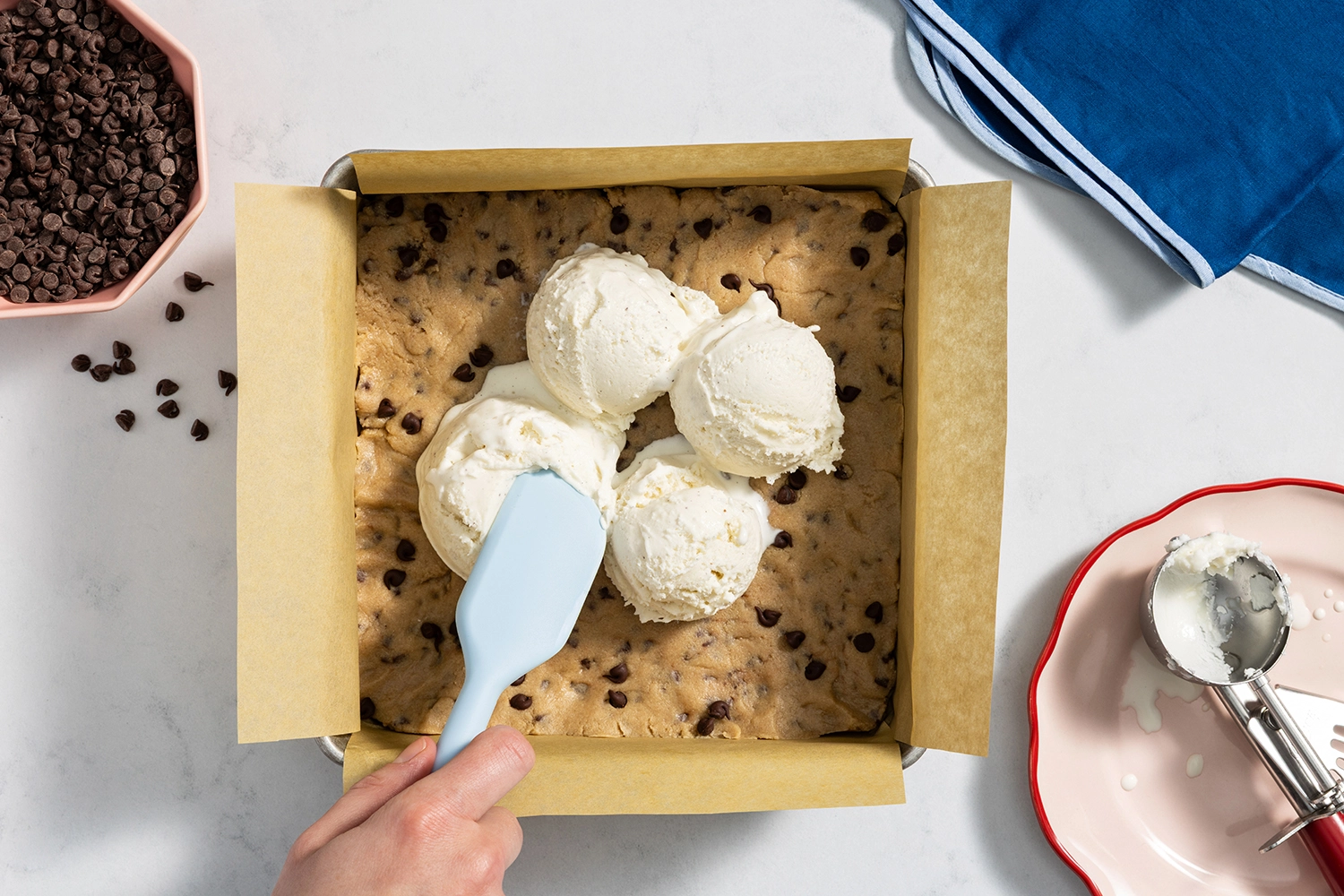 Spreading vanilla ice cream over chocolate chip cookie dough in a lined baking pan.