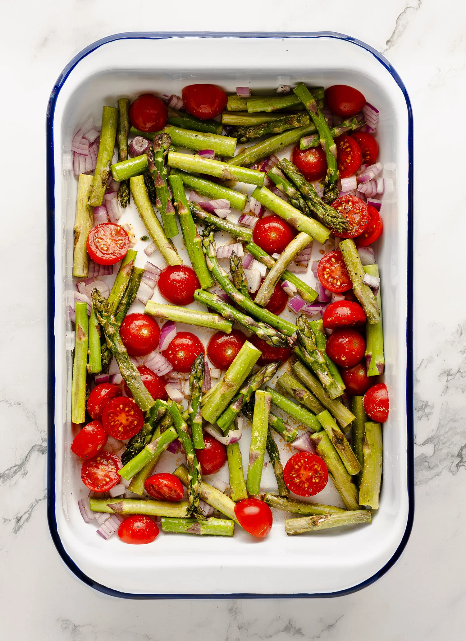 Seasoned asparagus, onions, and tomatoes with olive oil in an enamel baking pan on a countertop.