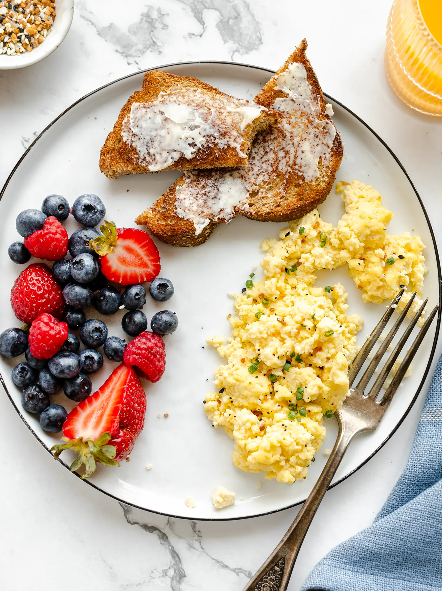 Plated cottage cheese scrambled eggs garnished with chives and everything but the bagel seasoning paired with toast, juice, and a side of fruit.