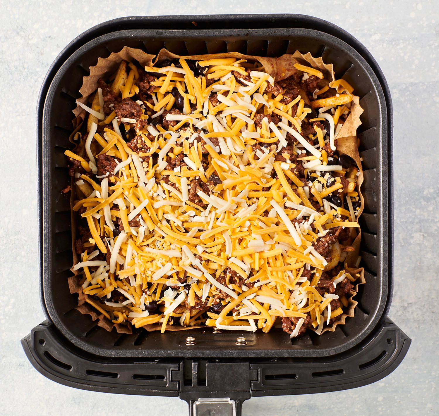 Fries in an air fryer topped with cooked ground beef and shredded Mexican cheese.